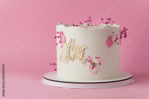 Elegant white birthday cake with pink blossoms and gold Happy Birthday topper on the pink background