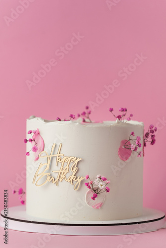 Elegant white birthday cake with pink blossoms and gold Happy Birthday topper on the pink background