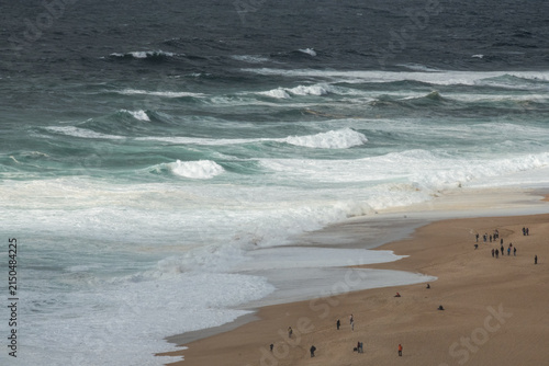 Aerial coastal view of sandy beach and powerful Atlantic waves near Nazare, Portugal. Scenic ocean shoreline with surf conditions, cliffs, and natural seaside landscape under cloudy skies.