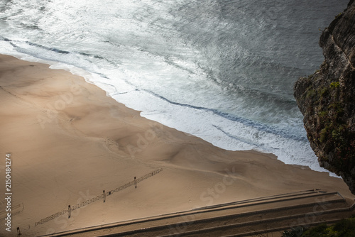 Dramatic aerial style view from high cliffs overlooking sandy beach and Atlantic Ocean waves in Nazaré Portugal beneath cloudy sky creating scenic coastal travel landscape scene.