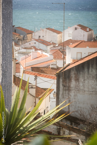 Scenic panoramic view of Nazare village in Portugal with white houses, red tiled roofs, green hillside and Atlantic Ocean coastline under dramatic clouds and bright daylight scenery.