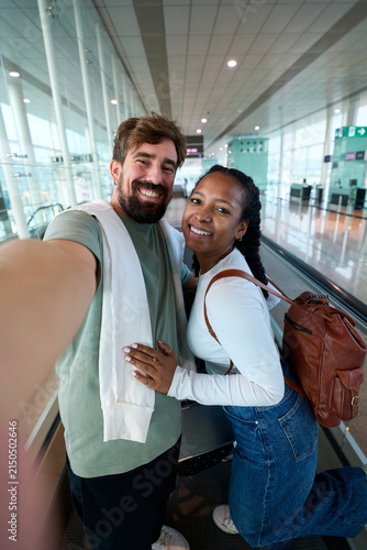 Young diverse couple smiling and taking a selfie on an airport moving walkway, looking at camera cheerfully. Vertical image.