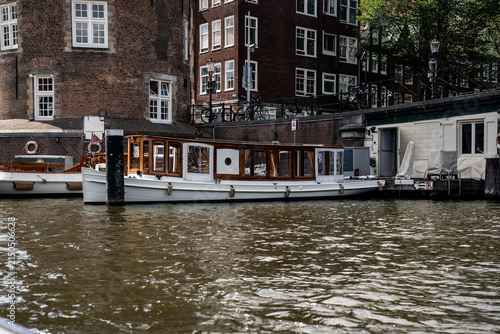 Classic wooden salon boat moored on the canal near the historic De Waag building in Amsterdam, Netherlands.