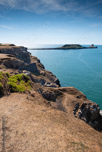 A sunlit view across drought-dried grassland and rugged cliffs on the Gower Peninsula, with the distinctive Worm’s Head rising beyond the sparkling Atlantic under a clear summer sky.