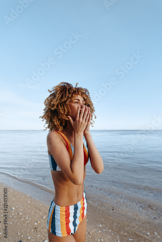 Photography Woman beach ocean curly hair covering mouth hands on sandy shore, striped swimsu