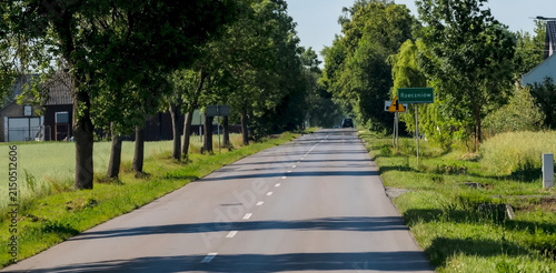  Droga asfaltowa w województwie mazowieckim, a konkretnie w pobliżu Rzeczniowa w Polsce. Droga jest otoczona drzewami i trawą, a nad nią rozciąga się czyste, błękitne niebo. Znak drogowy