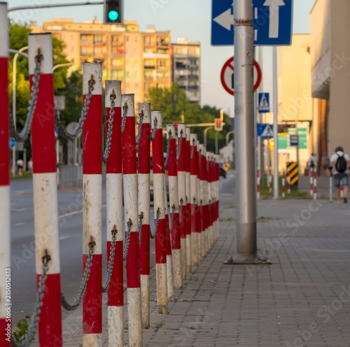 Czerwono-białe metalowe słupki bezpieczeństwa drogowego wyznaczają chodnik w Ostrowcu Świętokrzyskim. Są połączone łańcuchami i biegną równolegle do ulicy. W tle widoczne są znaki drogowe i budynek 