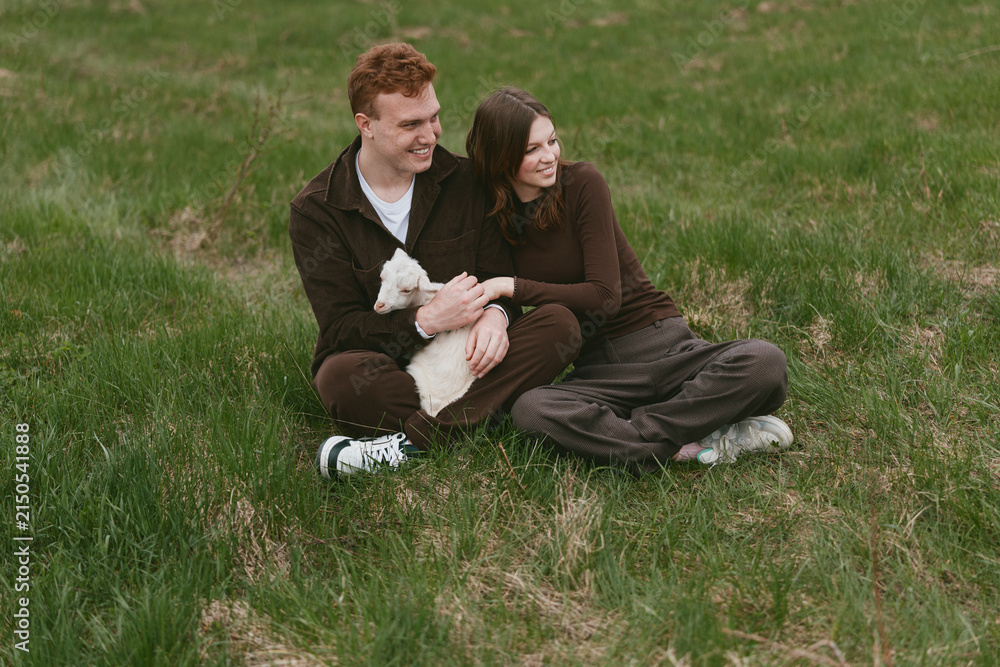 Obraz premium Couple outdoors, relaxed on a grassy field with a friendly goat kid nearby, smiling and affectionate, casual clothing, warm sunlight, gentle breeze, natural colors, peaceful moment, nature lifestyle