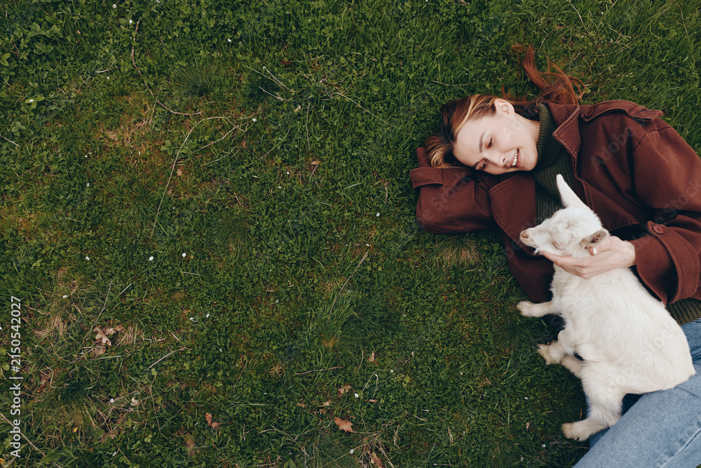 Obraz premium Relaxed woman lying on grass with goat kid, enjoying a peaceful outdoor moment, casual attire, sunlight, friendship, gentle bond between human and animal, serene scene, happiness, natural setting,
