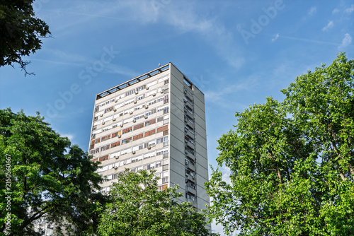 Photography A tall brutalist residential block rises above trees in Novi Beograd, Belgrade, sserbia, showing the facade, concrete massing and functional design associated with socialist-era urban housing