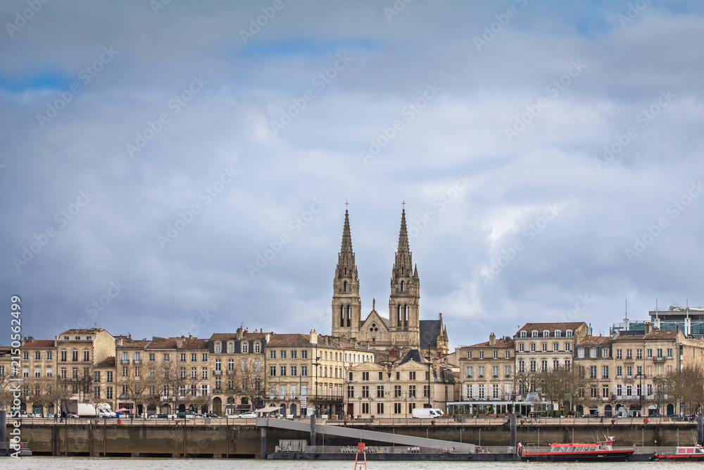 Fototapeta premium Panoramic view of the Garonne quays in Bordeaux, France, with historic waterfront buildings and Saint Andre Cathedral rising behind the cityscape during a winter afternoon under a partly cloudy sky.