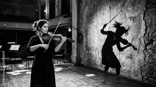 Young woman plays violin on stage, shadow figure bows intensely on wall