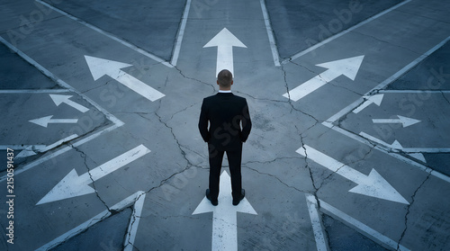 A man in a black suit stands at a crossroads with multiple white arrows painted on a cracked, gray asphalt surface, facing forward featuring businessman, formal, direction, decision and choice