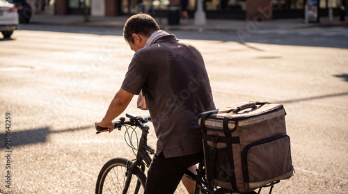 Delivery Cyclist's Sweaty Ride: A lone delivery cyclist, his back showing sweat stains under the summer sun, pedals a bicycle loaded with a large insulated delivery bag through a sunlit urban street.