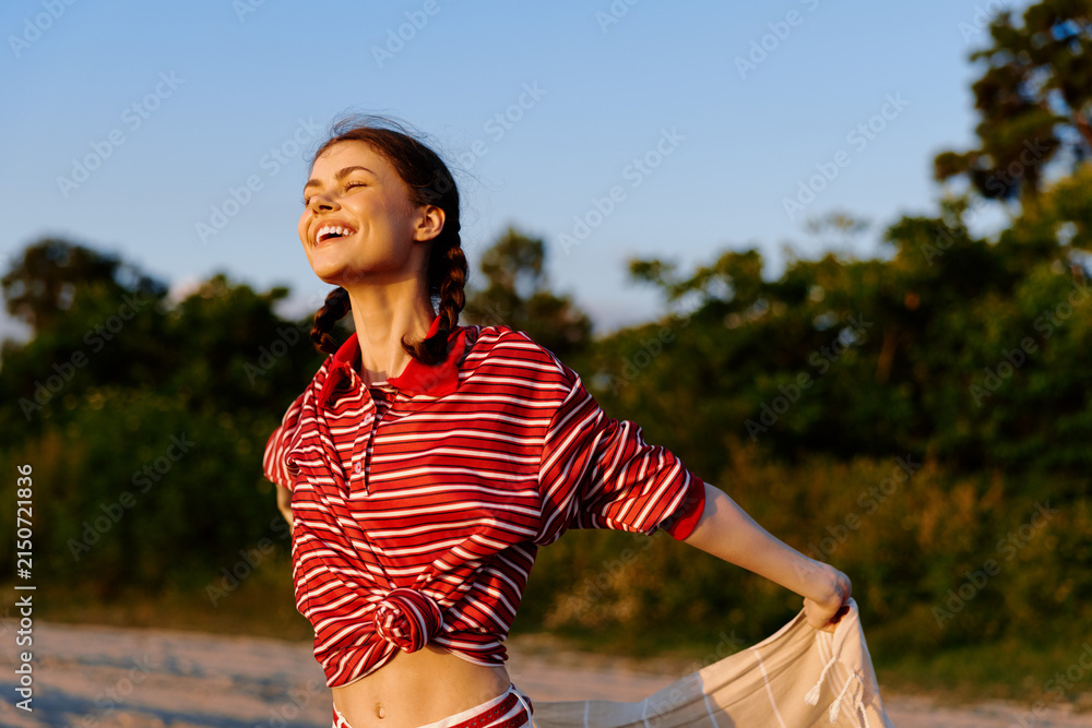 Obraz premium A smiling woman in a red striped shirt enjoys the outdoors, arms open, breeze in her hair. A bright natural setting conveys wellness, confidence, and carefree lifestyle.