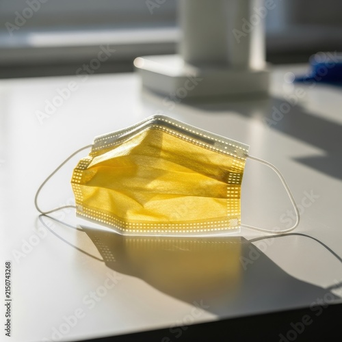 A bright yellow disposable medical face mask rests on a white surface with soft lighting and distinct shadows, emphasizing its protective qualities.