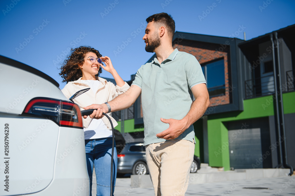 Fototapeta premium Couple charging electric car at home near modern house