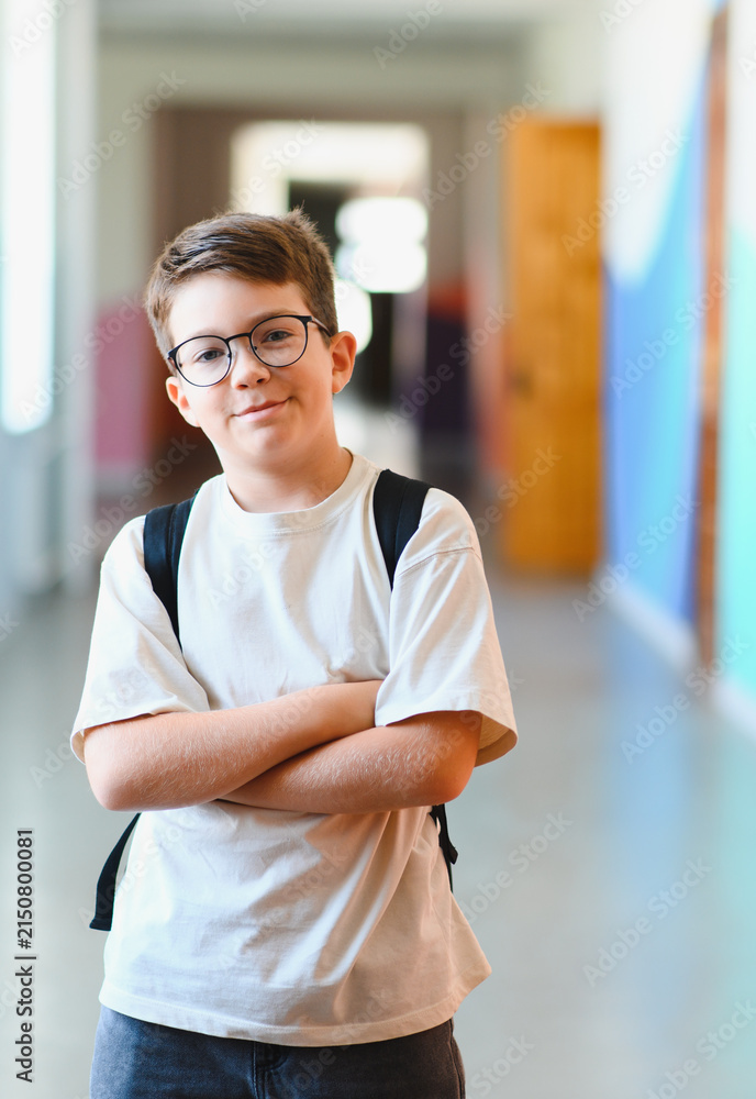 Obraz premium Smiling pupil wearing eyeglasses and backpack crossing arms in school hallway