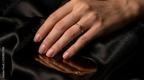 Elegant female hand wearing a gold diamond ring resting on luxury black silk with reflection.