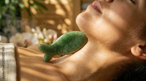 Woman receiving relaxed facial spa massage with a green jade fish-shaped gua sha tool on neck and jawline.