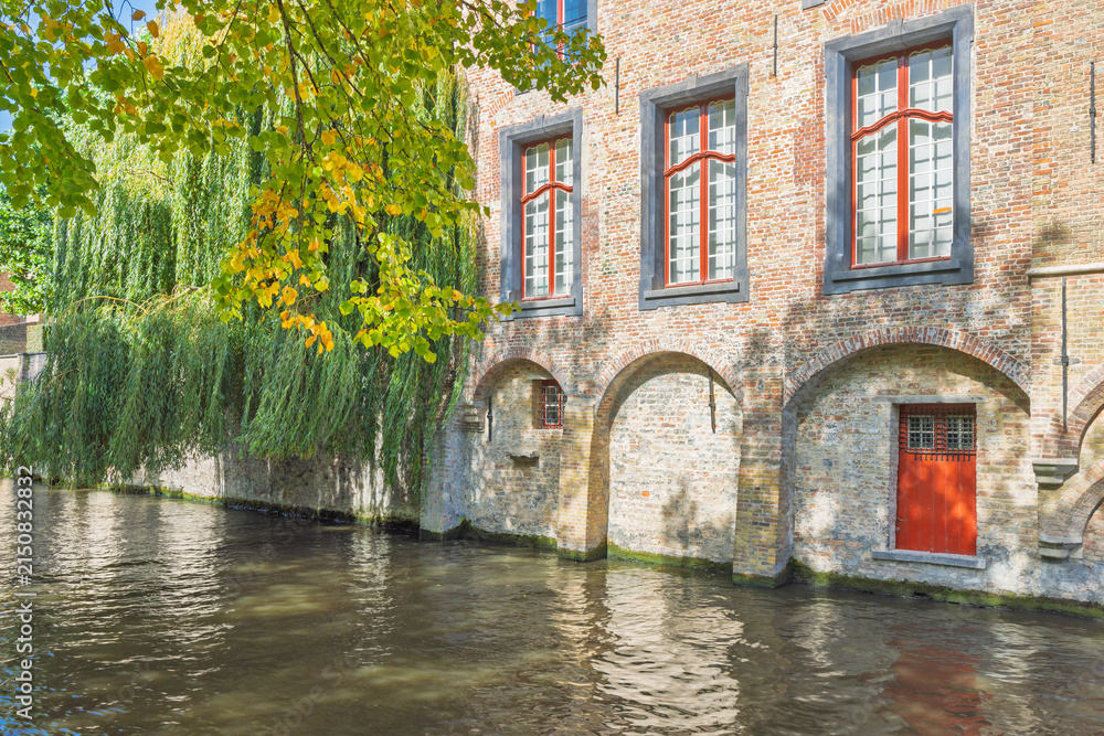 Obraz premium Bruges town in Belgium, Europe. Historic brick building with arched foundations along a serene canal, lush green foliage cascades over the water, under warm sunlight