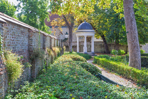 Bruges town in Belgium, Europe. Charming garden path winds past a historic stone wall, lush greenery, and a delightful classical gazebo under bright sunlight in an old town setting
