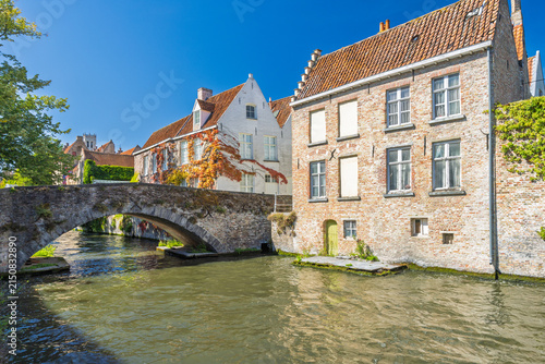 Bruges town in Belgium, Europe. Historic European Canal Scene with Ancient Stone Bridge Arching Over Tranquil Water, Flanked by Traditional Brick Buildings Under Clear Blue Sky