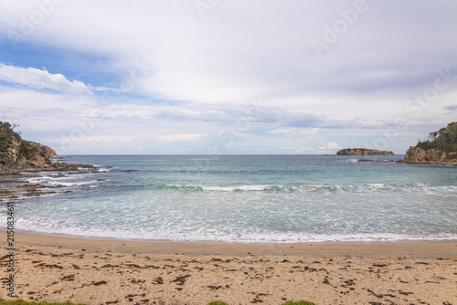 A lovely cove beach called McKenzies Beach is surrounded by rocky, vegetated headlands and an island in a turquoise sea on a calm and overcast day at Batemans Bay in New South Wales, Australia.