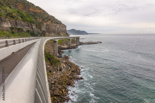 The Ocean bridge road on Grand Pacific Drive curves on its pillar supports far above a rocky shore between the sea and a steep cliff on a calm, overcast day at Coalcliff in New South Wales, Australia.