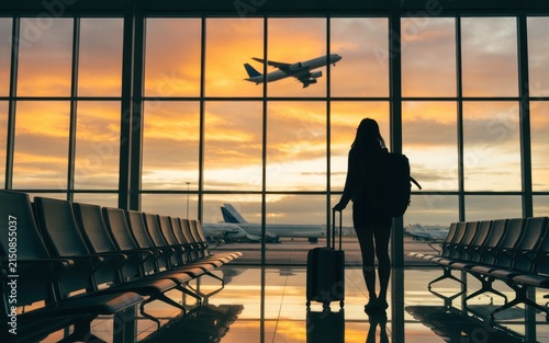 Silhouette of a traveler with a suitcase and backpack standing in an airport terminal looking at an airplane taking off during a golden sunset through large glass windows