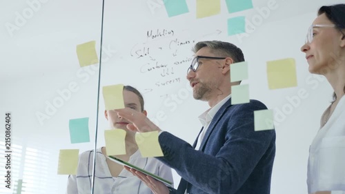 Business team collaboratively brainstorming new strategies and ideas on a glass whiteboard, creating a project plan with sticky notes and marker in a modern office meeting