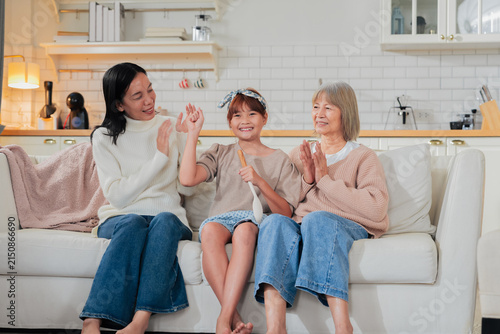 Obraz na plátne A little girl sings into a wooden spoon microphone while her mother dances and grandmother claps along on the sofa a playful, joyful family moment of impromptu music and shared laughter at home