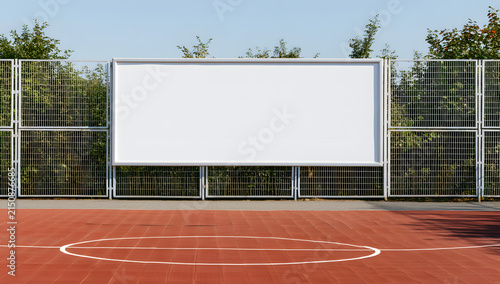 A basketball court outdoors has a white billboard affixed to a metal fence, used for signage or advertising