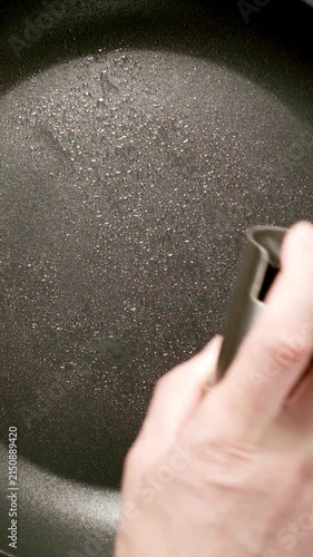 TOP VIEW: Man's hand sprays vegetable oil from dispenser (spray gun) on a black frying pan on stove