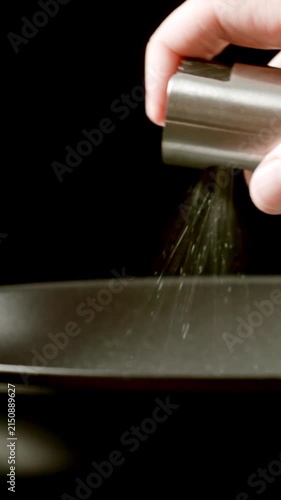 Man's hand sprays vegetable oil from dispenser (spray gun) on a black frying pan on stove