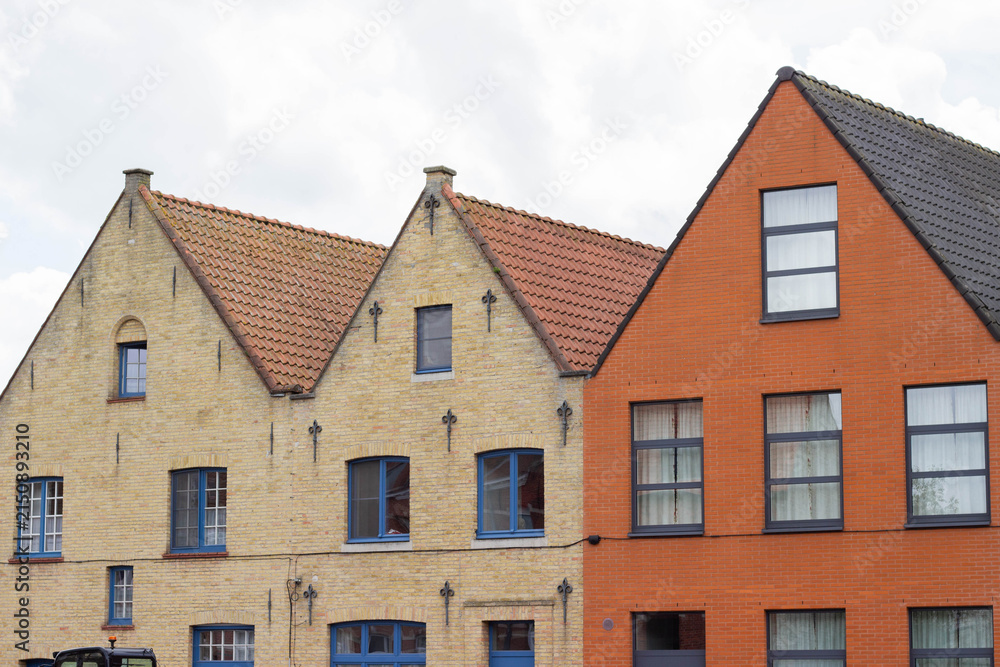 Naklejka premium Historic brick and stone buildings with gabled roofs in a row, featuring blue windows and a mix of red and yellow facades against a cloudy sky