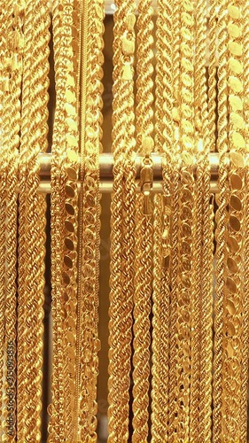 A tight closeup shows multiple gold necklaces and chain styles hanging vertically on a display bar, gleaming under bright light with subtle movement and sparkling reflections