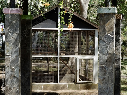 Фотография Park birdhouse cage framed by stone pillars with hanging orange flowers and green trees in daylight