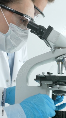 Scientist wearing protective mask, safety glasses, and gloves, examining a slide under a microscope, conducting important medical research and experimental analysis in a laboratory