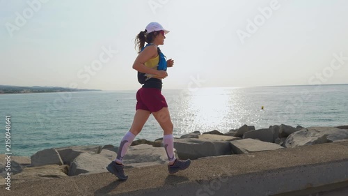 Woman jogs along a concrete coastal path. She wears a white cap and maroon shorts. Sunlight glints off the sea water. Her expression shows focus. This footage captures active moments by the sea