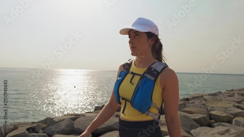 Sunlight glints off the sea. Woman wears life vest and white cap. She stands calmly on rocky shore. Her posture shows readiness for activity. Ideal for safety training or adventure content