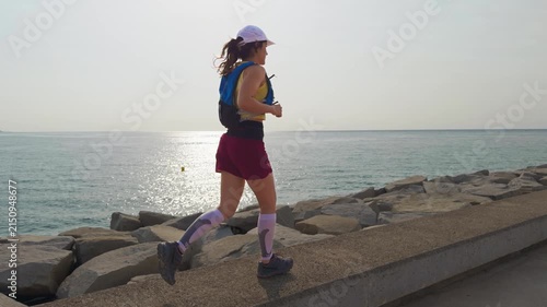 Woman runs steadily along a coastal path. She wears a white cap and athletic gear. The sea glows under the setting sun. Her expression shows focus and energy