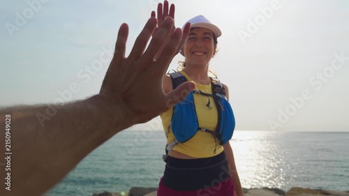 Woman smiles by the sea. She wears a life vest and white cap. Man's hand reaches toward her. She looks happy and relaxed. Sunlight glints on the water