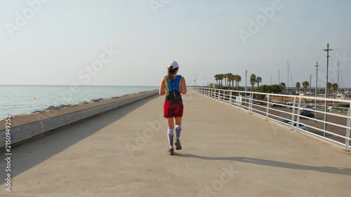 Sunlight glimmers on the ocean boardwalk. Woman jogs forward in red shorts and blue vest. Palm trees line the coastal path. Her focused stride blends with calm seas