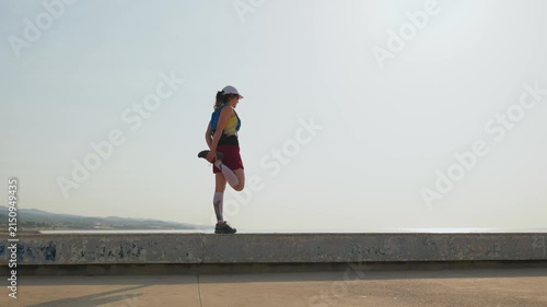 Girl stretches on seaside ledge. Clear sky and distant hills frame scene. She wears bright top and red shorts. Focused expression shows determination. Ideal for fitness content, wellness blogs