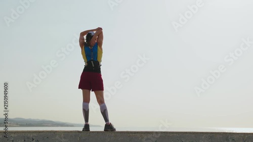 Woman stretches arms above head on sandy beach. Focused posture shows determination. Early morning light illuminates calm sea and hills. Close-up reveals tired yet resilient expression