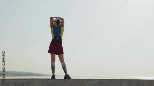 Woman stretches on a sunlit beach. She wears a blue vest and maroon skirt. Her arms are raised, showing calm focus. The ocean stretches behind her, hazy and serene