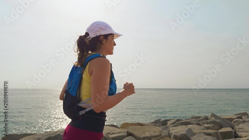 Woman runs along rocky coastline at sunset. She smiles, enjoying ocean breezes. Sunlight glints off the sea behind her. Her bright athletic outfit stands out. Ideal for fitness or travel content