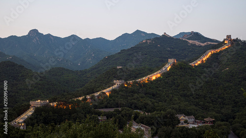 Foto The Great Wall of China stretches across a rugged, forested mountain landscape at dusk, with sections of the ancient fortification illuminated by warm golden lights