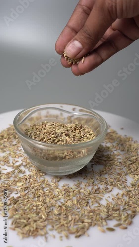 A close up view of a hand drop seasoning into the glass bowl. A glass bowl is full of a seasoning like Fennel seed on the table. Fennel seeds on a white background